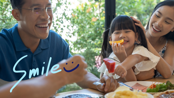 左側に”Smile”と記載。草木の見える窓際で料理を食べながら笑っている幼女を挟むように両親が並んで座った画像
