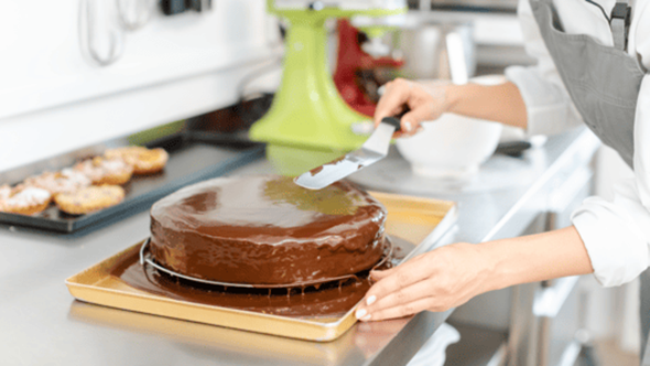 調理室の調理台でチョコレートのホールケーキにチョコをコーティングしている画像