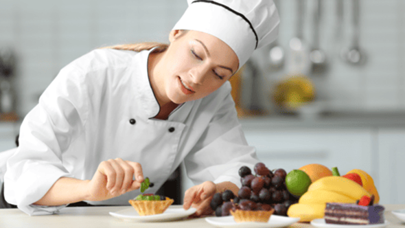 調理室の調理台でコック帽にコックシャツを着た女性がタルトに果物を乗せようとしている画像